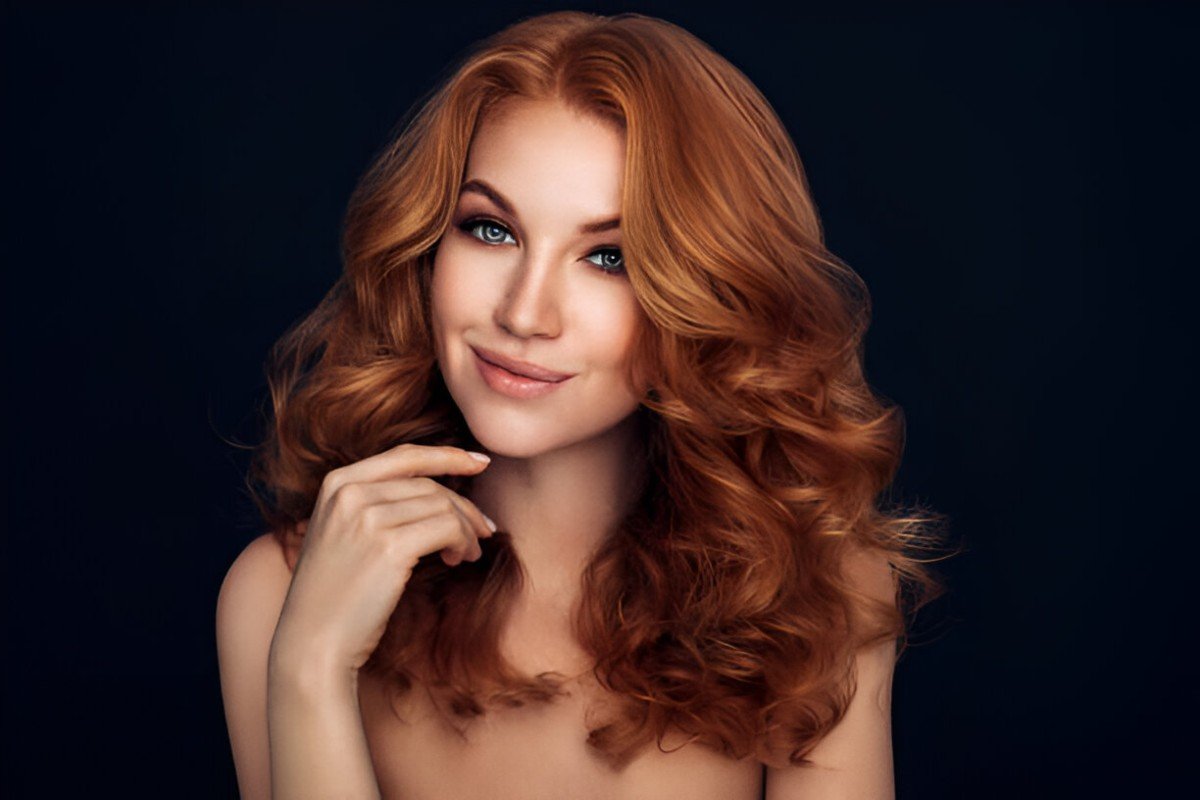Portrait of a smiling woman with voluminous red curls, flawless skin, and subtle makeup against a dark background.